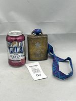 Photo of the bronze-tone Olympic cowbell next to a soda can for size reference, showing the colorful woven lanyard and metal body with patina.