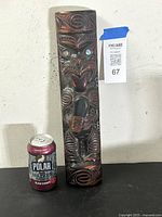 Front view of a 15-inch tall wooden Maori tekoteko figure with carved traditional motifs and paua shell inlaid eyes next to a can for scale.