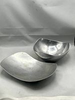 Two Nambe cast aluminum serving pieces, a triangular bowl and freeform platter, with visible surface wear and scratches.