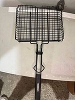 Top-down view of rectangular wire mesh BBQ grill basket with folding handle and locking clasp, placed on a white table.