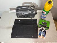 Electric tire pump, two black license plate holders, 150W power inverter box, Turtle Wax carpet cleaner spray with attached brush, and pack of 2 Sylvania SilverStar light bulbs arranged on table.
