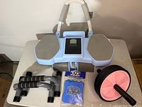 Photo showing an ab roller, gray push up handles, black lifting grips, and blue workout gloves in packaging, arranged on a flat surface.