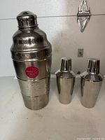 Front view of one extra large hammered martini shaker with red 'Martini & Rossi' label, and two smaller smooth shakers side by side