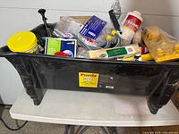 Top view of the large black Purdy painters bin filled with assorted tools, plumbing parts, sanding compounds, and small hardware boxes.
