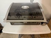 Closed portable Cuisinart BBQ grill showing branded lid with temperature gauge and carry handle.