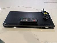 Front view of the black Sony 3D BluRay DVD player with remote control and power cable on top, showing disc slot and buttons.