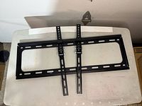 Photo shows black metal TV wall mount brackets laid flat, consisting of a rectangular mounting frame and two vertical arms with adjustment holes.