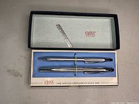 Open box showing two silver-tone Cross pens nestled in blue velvet inset, with original branded packaging lid.