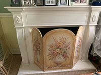 Front view of white decorative fireplace mantel with floral embellishments and the yellow floral three-panel decorative screen positioned inside the fireplace opening.