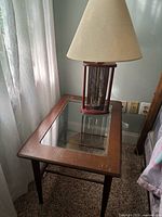 Photo of a rectangular end table with wooden frame and glass top, standing on four legs next to a window with sheer curtains. On the table sits a lamp with a wooden and glass base and beige fabric lampshade.