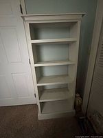 Photo of a narrow wooden book shelf with four shelves and a decorative top edge, painted light color and showing some wear marks.