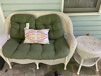 White wicker loveseat with green cushions and one multicolor decorative pillow along with one round wicker side table