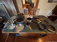 Overview of all pots and pans on green countertop, showing variety of sizes and materials including cast iron, stainless steel, and non-stick surfaces