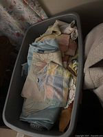 Plastic bin filled with assorted folded table linens in pastel and earthy colors, including napkins, table runners, and small fabric pieces.