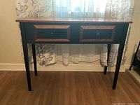 Front view of tall narrow table with two drawers, wood-finish top, black legs and frame.