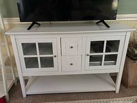 White wooden TV stand with two glass door cabinets, two central drawers, and an open shelf at the bottom, supporting a TV on top.
