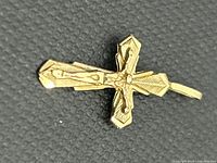 Close-up photo of a 14k yellow gold cross pendant showing intricate raised detailing and textured geometric patterns on a black textured background.