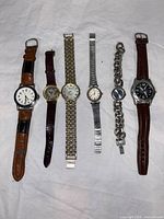 Photo showing six assorted wristwatches lying side by side on white background, arranged in a single row with various bands and dial designs visible.