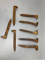 Eight rusty antique railroad spikes displayed on a light surface, showing their varied shapes and condition.