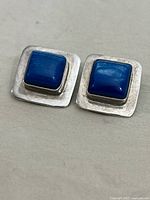 Top view of a pair of 925 silver clip earrings, square shape with synthetic blue tiger's eye stones set in the center.