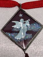 Front view of the square boxed pendant with silver angel playing trumpet, blue glittery background, silver star, and red hanging Swarovski crystal teardrop on red ribbon.