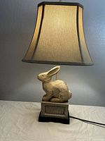 Full view of the table lamp showing beige square-tapered lampshade and distressed resin rabbit on rectangular crackle finish base with black platform.