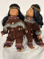 Two Indigenous dolls with porcelain heads, hands, and feet dressed in detailed brown fringed and beaded clothing, black hair, and patterned headbands posed together
