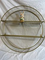 Front view of large round gold metal grate wall shelf with two horizontal shelves, with porcelain bunny figurine placed on the top shelf.