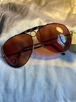 Front angled view of sunglasses showing rose amber tinted lenses, gold metal frame with tortoiseshell detail on nose bridge and arms, with original tags attached.