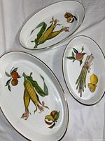 Set of three oval porcelain dishes with vegetable and fruit paintings and gold rims photographed on white fabric.