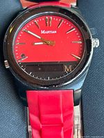 Close-up of Martian Notifier smartwatch with red analog dial, digital display, and red segmented silicone band on a clear surface.