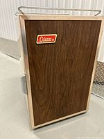 Front view of the vintage Coleman ice chest featuring a wood grain panel with a Coleman logo and metal framing.