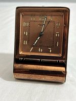 Front view of the square copper travel alarm clock folded closed, showing the clock face and casing.
