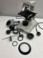 Wide view showing microscope base, mechanical stage, objective lenses, two eyepiece assemblies, and individual lens adapters and parts spread out.
