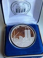 Collectible coin in case showing full coin design with skyline and Twin Towers, inscription 'We Will Never Forget', and 'One Dollar'.