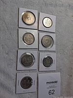 12 world coins displayed individually in white cardboard holders, showing obverse sides with various inscriptions and imagery.