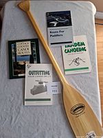 The wooden Feather Brand Caviness Woodworking canoe paddle laid flat alongside five canoeing and outdoor books arranged neatly on a white surface.