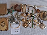 Full overview of mixed costume jewelry and accessories lot laid on white fabric. Items include bracelets, rings, brooches, a wristwatch with white strap, and a small gold-tone purse or wallet.