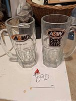 Two large A&W glass mugs side by side on a surface. One mug has the 'A&W Since 1956' logo, the other shows the 'A&W Root Beer' logo.