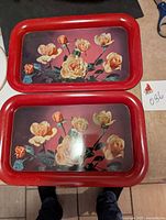 Both metal trays with red borders and cream and peach rose prints side by side on carpet.