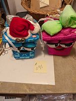 Two plush holiday presents, one blue in snowflake print with Santa hat and two jingle bells, and one pink with green bow, both with facial features.