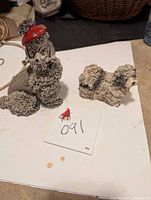 Two ceramic spaghetti dog figurines, one larger with red hat and detailed curly fur, the other smaller and simpler, placed on a white surface with tag 091.
