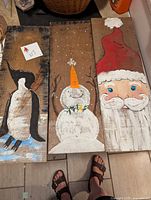 Three Christmas-themed paintings on barn board depicting a penguin, snowman, and Santa Claus.