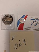 Coin with old Greyhound bus design in plastic case and 75th anniversary pin on card with some writing in background.
