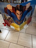 Front view of Superman cardboard display showing Superman's face and fists flying forward with cityscape background and product logos below.