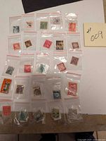 Overall view showing 25 individual stamps, each in small plastic bags, arranged on a flat surface with a paper label showing #009
