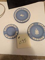 Photo showing three small Wedgwood blue jasperware plates and two ashtrays arranged on a white surface, highlighting the classic white leaf relief designs and central royal crests on plates.