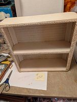 Front view of white wicker shelf showing two compartments and woven wicker frame.