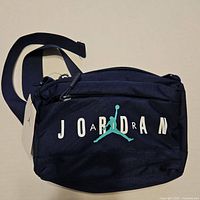 Front view of black Air Jordan fanny pack showing white AIR JORDAN lettering and teal jumpman logo, with adjustable strap and attached tag.