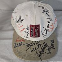 Front and brim view of the white PGA Tour baseball cap showing embroidered logo and multiple autographs in black, blue, red, and orange ink.